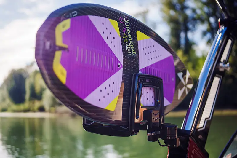 A close-up of the Flytrap Board Rack securely holding a brightly colored wakeboard with a purple and yellow design. The rack is mounted on a boat, with a scenic lake and trees in the blurred background.