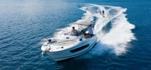High-angle shot of a white motorboat towing a wakeboarder across vibrant blue open water, leaving a curved white wake behind.