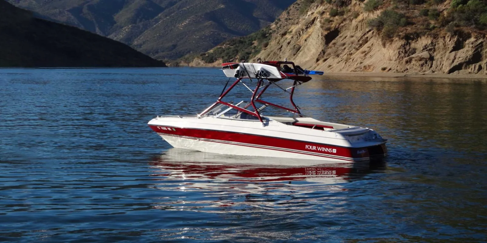 A red-and-white powerboat with a wakeboard tower and board racks idling on a scenic lake bordered by desert hills.